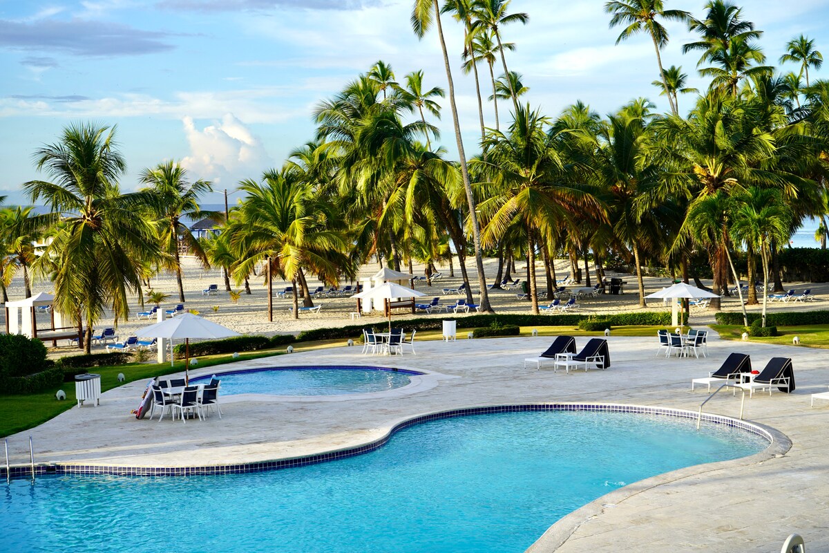 The outdoor pool area features two swimming pools, surrounded by lounge chairs and umbrellas. Lush palm trees provide shade, while the beach and ocean can be seen in the background, creating a serene atmosphere for relaxation.
