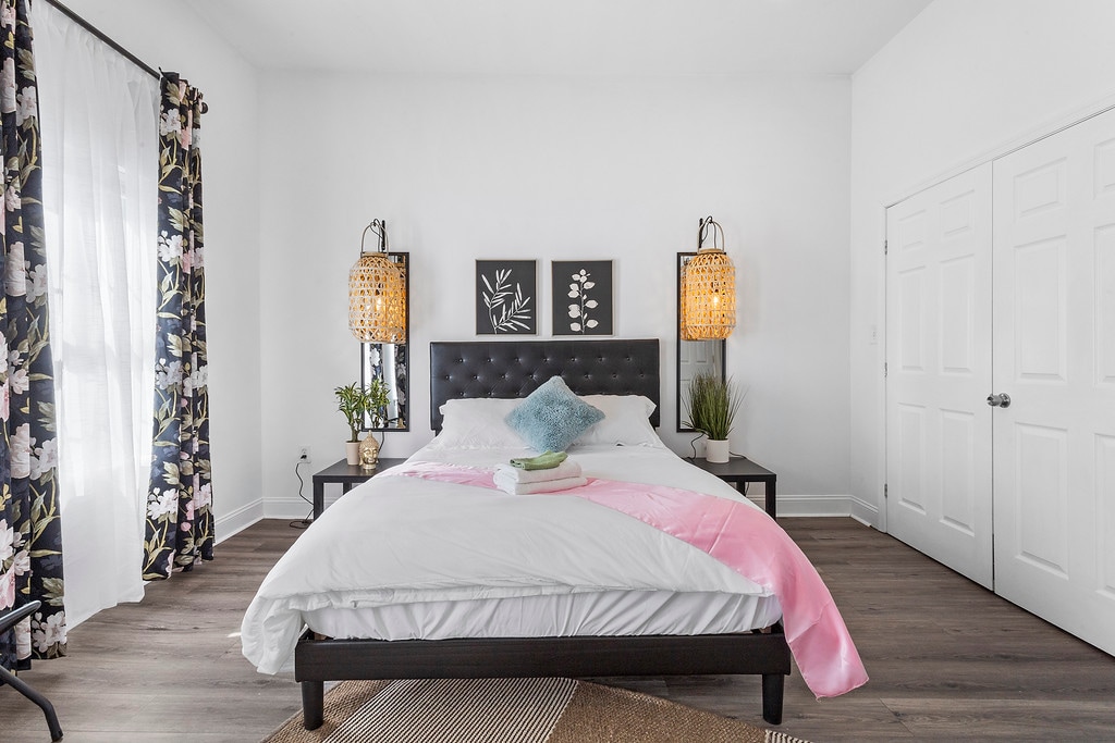 A bright bedroom features a spacious bed with a white comforter and decorative pillows. Two woven light fixtures flank the bed, casting a soft glow. The room is adorned with floral curtains and wall art, complemented by greenery in decorative pots. A large closet is visible.