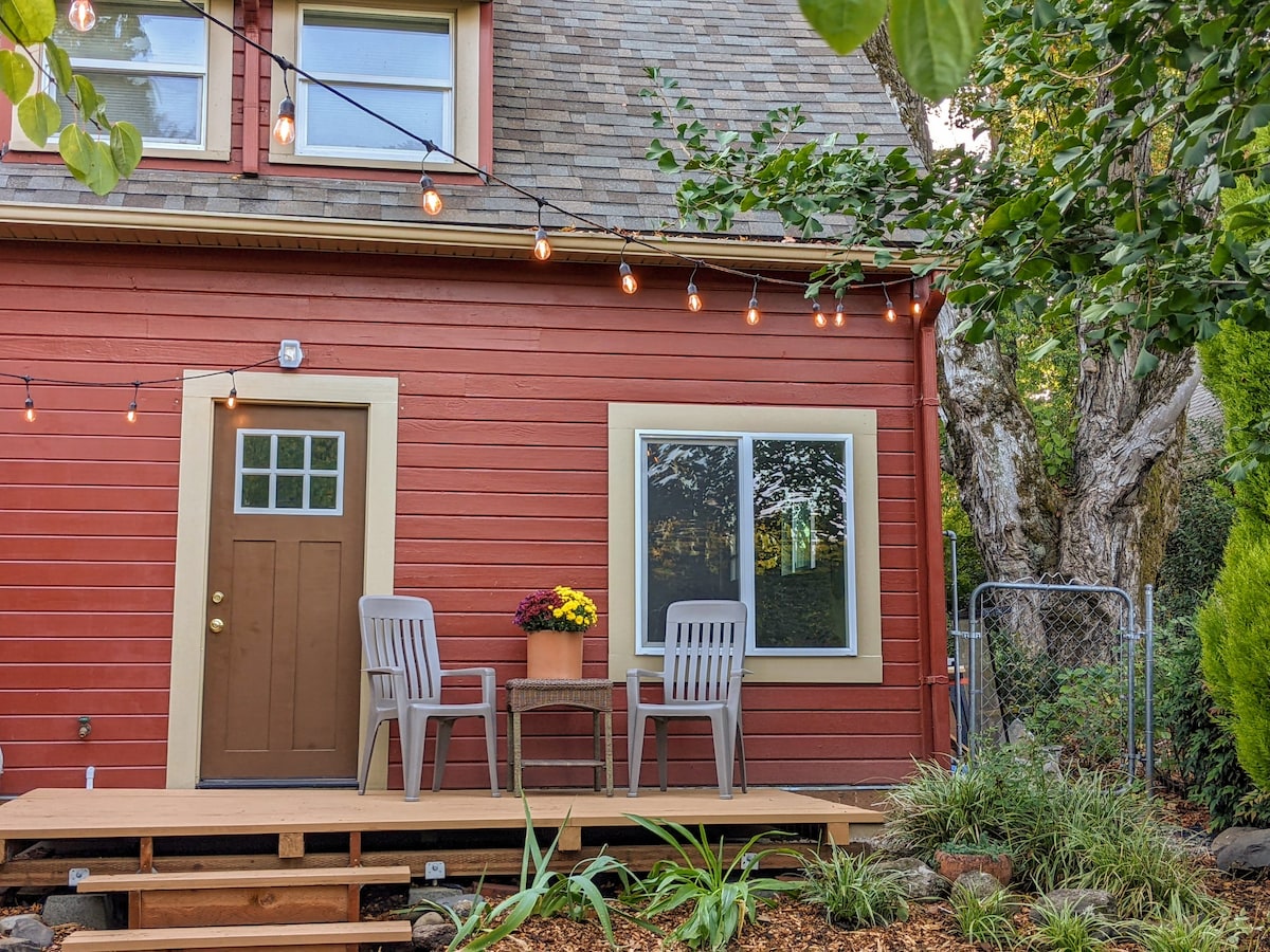 The exterior features a vibrant red wooden facade with a welcoming front door. Two gray chairs and a small table are positioned on a wooden deck, adorned with flowers. Warm string lights hang above, providing a charming ambiance in a garden setting.