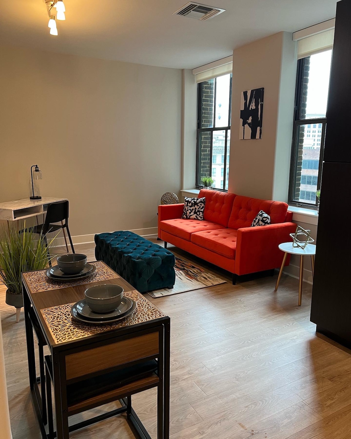 A modern living space is furnished with an orange sofa and a teal ottoman, creating a vibrant atmosphere. Natural light enters through two large windows, highlighting a small dining area with a stylish table and chairs. Decorative elements include plants and wall art.