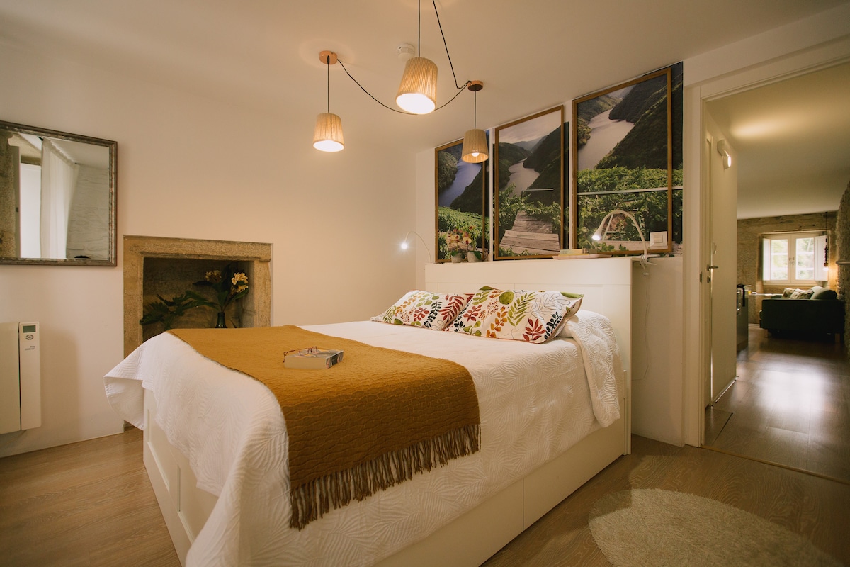 A bright bedroom features a spacious bed with decorative pillows and a warm throw. Pendant lights hang above, complementing the neutral decor. A mirror and framed artwork adorn the walls, with a glimpse of the living area visible through an open doorway.