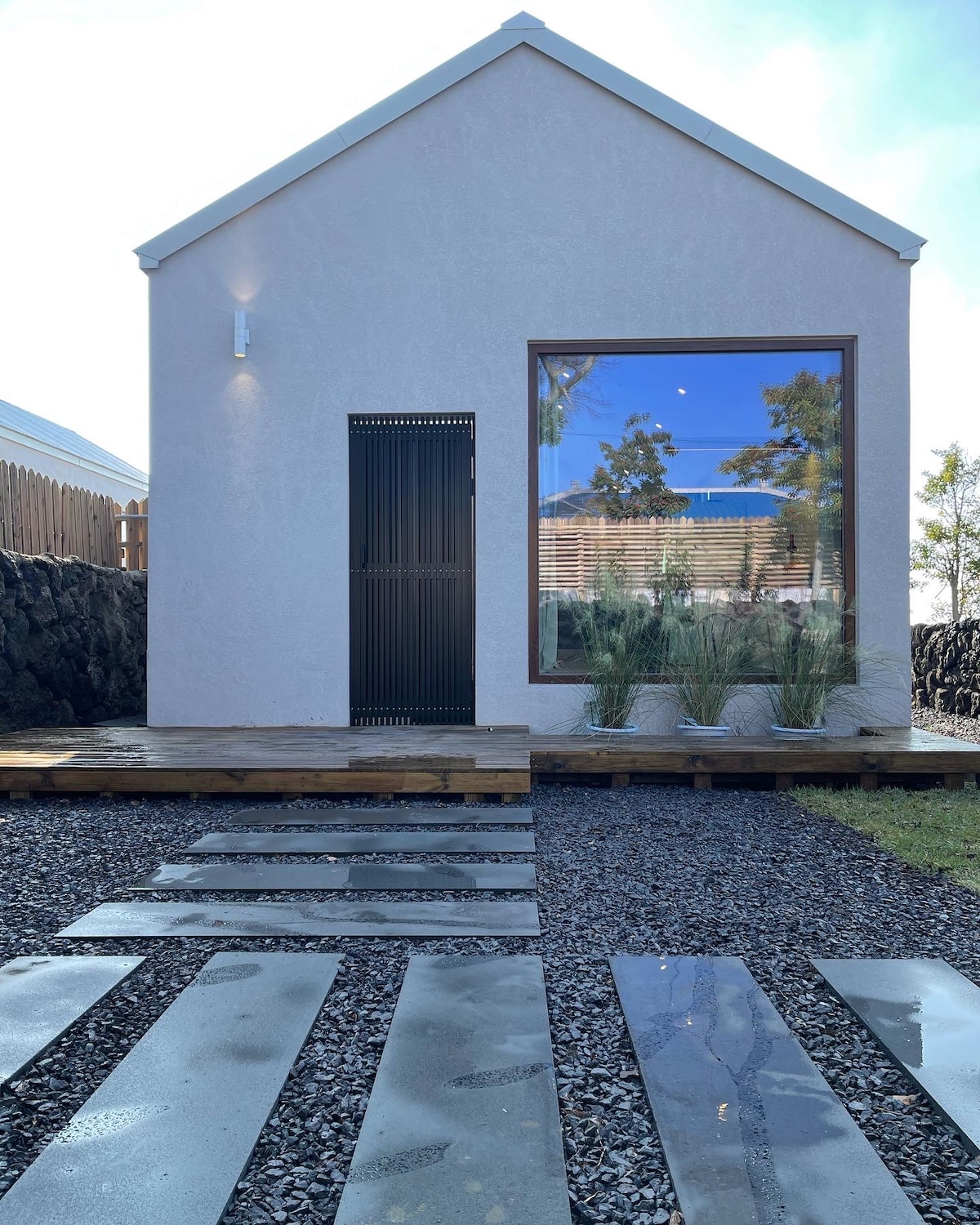 The exterior of a minimalist building is shown, featuring a large window and a sleek entrance door. A pathway of stepping stones leads through a gravel area, complemented by potted plants near the entrance, set against a clear sky.