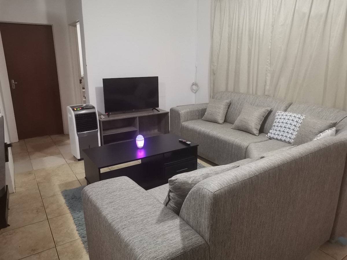 A lounge area features a spacious gray sectional sofa arranged around a dark coffee table. A television is positioned on a stand, complemented by an air conditioning unit nearby. Natural light filters through sheer curtains, creating a bright and comfortable environment.