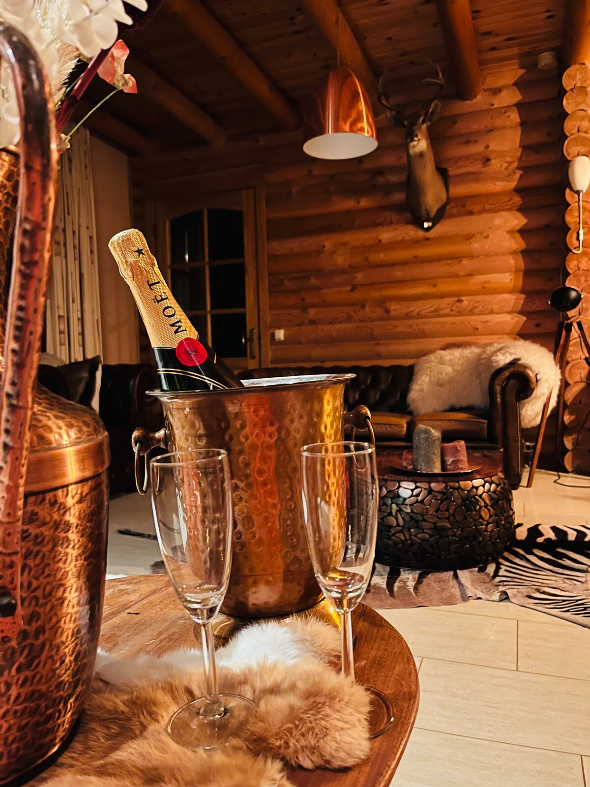 A copper ice bucket holds a bottle of champagne, surrounded by two champagne flutes. The rustic interior features a wooden log wall, a leather sofa, and a plush rug, creating a cozy atmosphere. Warm lighting from pendant fixtures enhances the inviting space.
