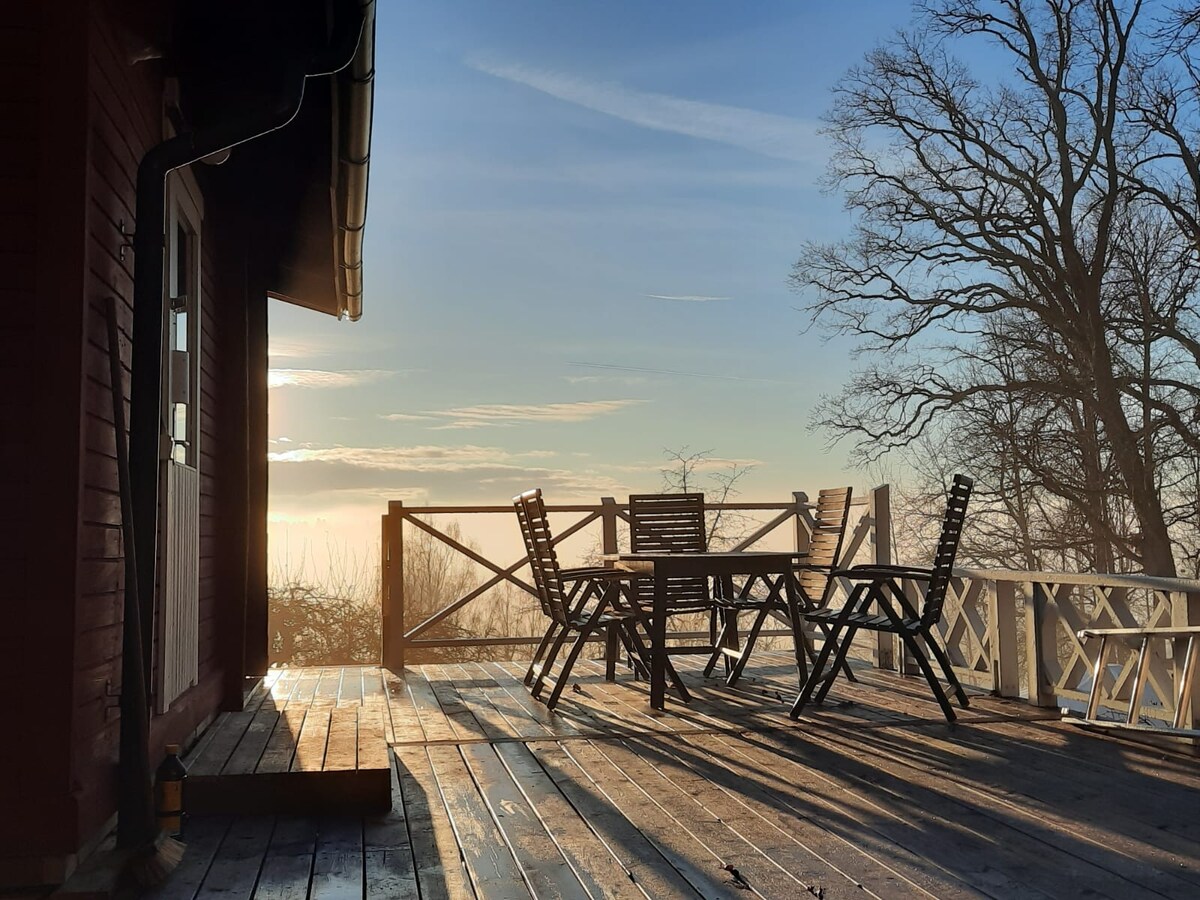 A wooden terrace offers a set of outdoor furniture positioned towards a scenic view, bathed in soft morning light. A railing surrounds the area, and the surrounding landscape is gently shrouded in mist, creating a serene and peaceful atmosphere.