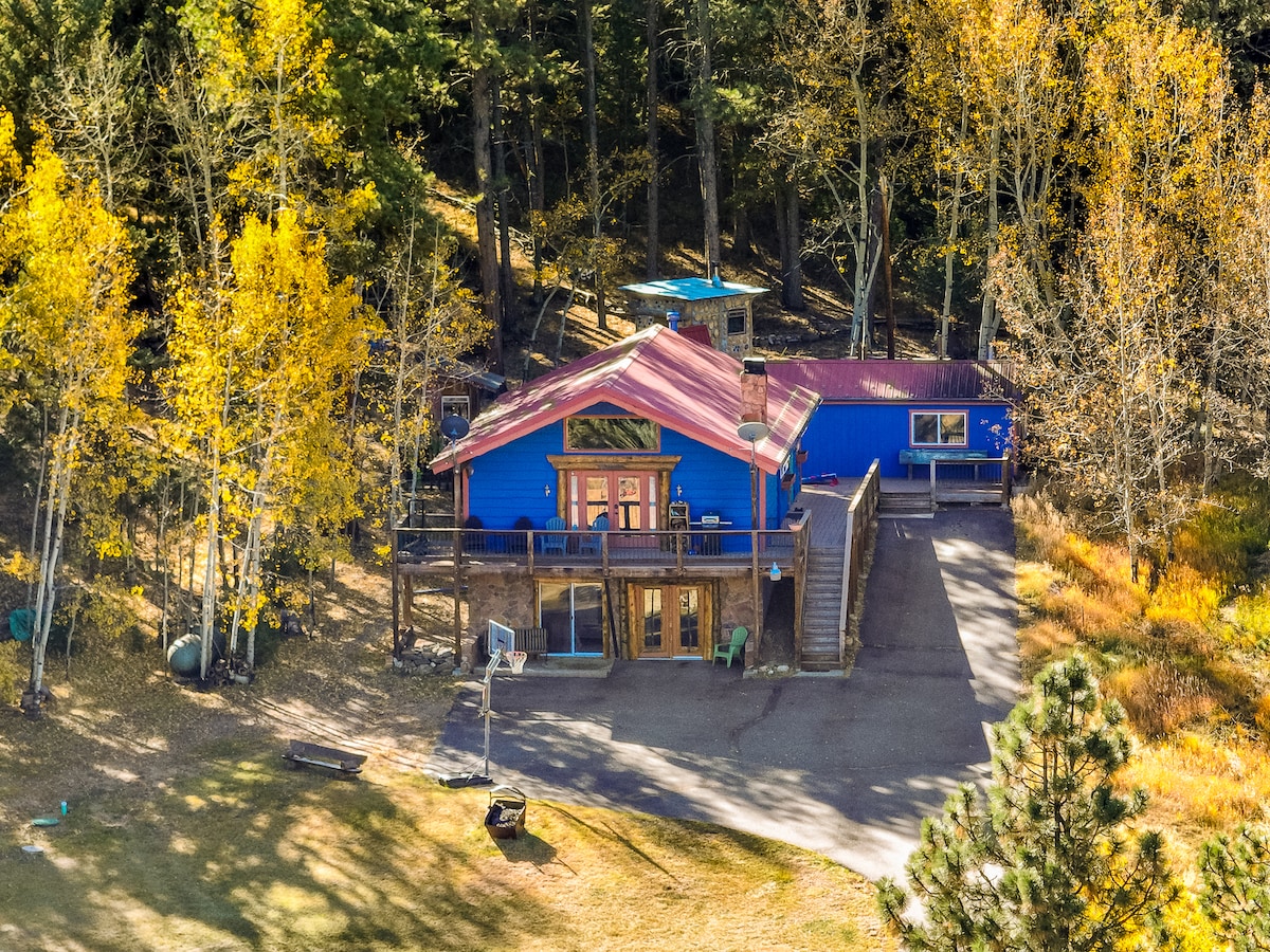 A colorful cabin is nestled among tall trees, showcasing a wraparound deck and a staircase leading to the entrance. Surrounding foliage features vibrant autumn colors, while a basketball hoop stands at the edge of a spacious yard, inviting outdoor activities.