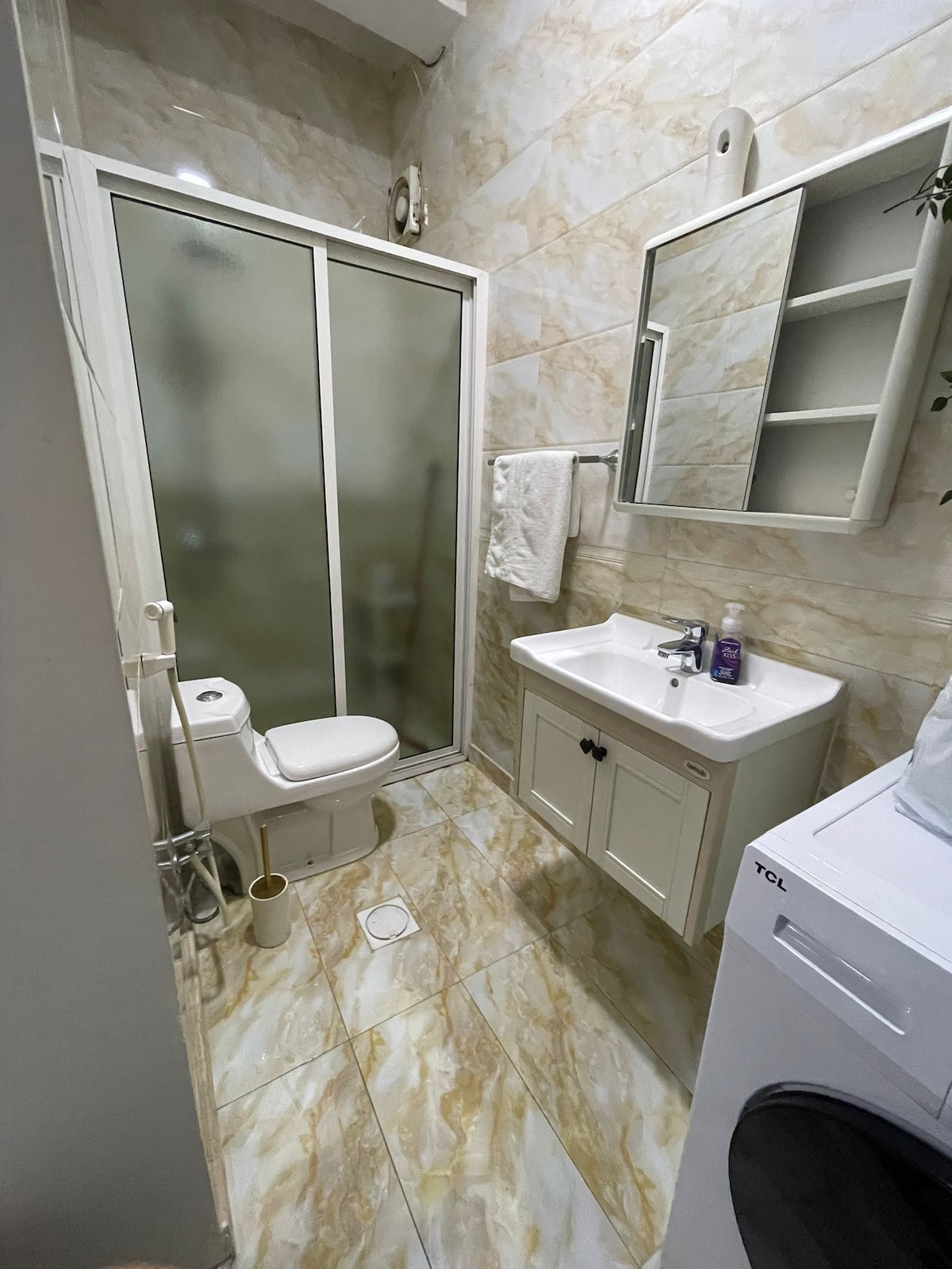 A compact bathroom features a glass shower enclosure and a white vanity with a sink. A toilet is positioned beside the vanity, and a wall-mounted mirror reflects the room's light. Shelves are visible above the sink, and a washing machine is located nearby.