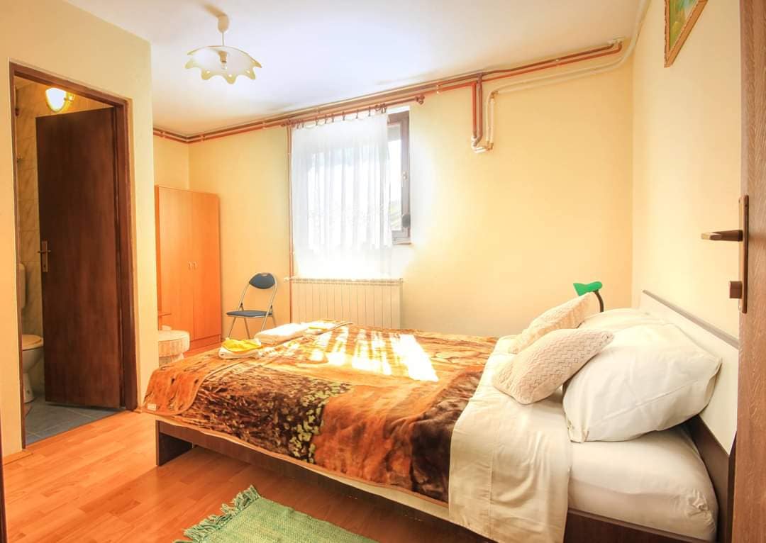 A cozy bedroom features a spacious bed dressed in warm bedding with decorative pillows. Natural light filters through a window adorned with sheer curtains, illuminating the light-colored walls. A chair is positioned beside the bed, and a wooden cabinet is visible in the background.