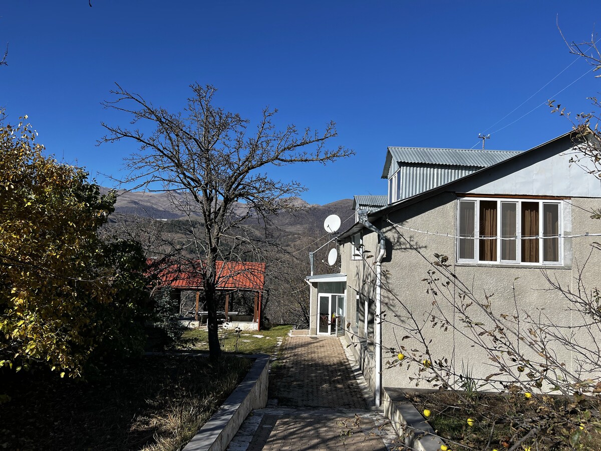 The two-story house is set against a backdrop of clear blue skies and distant mountains. A pathway leads to the entrance, flanked by trees and outdoor spaces, including a separate structure with a red roof. Satellite dishes are mounted on the building's exterior.