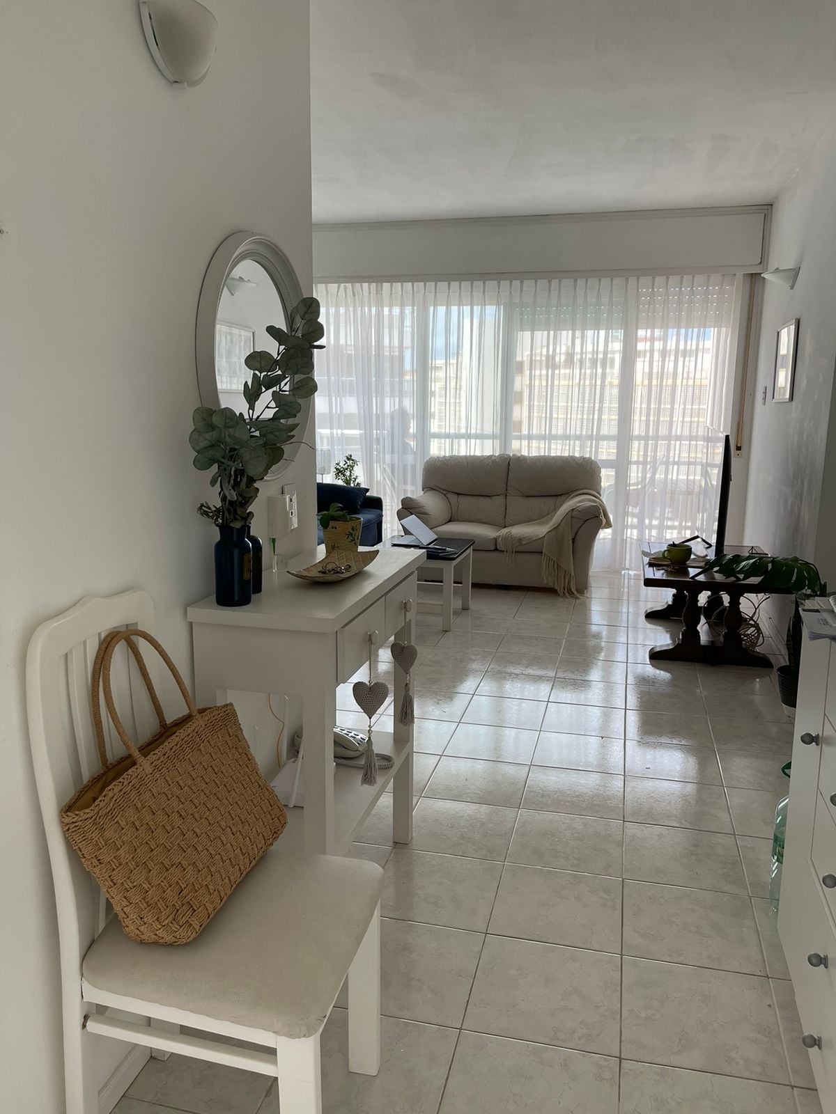 A bright corridor is lined with white tiled flooring, leading to a spacious living area illuminated by natural light through sheer curtains. A small table holds decorative items, while a chair with a woven bag rests nearby. A comfortable sofa is situated at the end of the corridor.