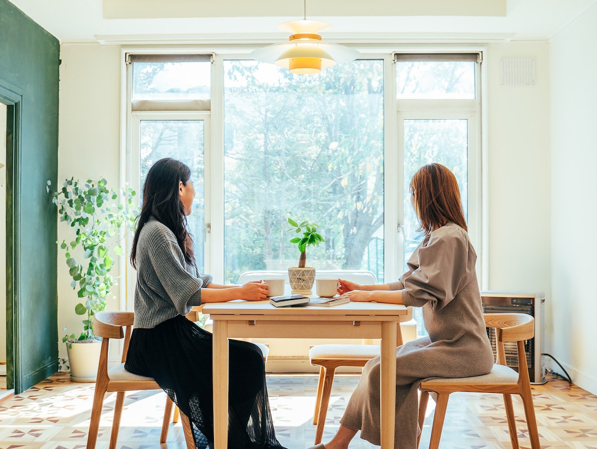 A light-filled dining area is highlighted by large windows overlooking greenery. A wooden table is flanked by two chairs, where two women are seated, engaged in conversation. A small potted plant sits at the center, enhancing the serene atmosphere.