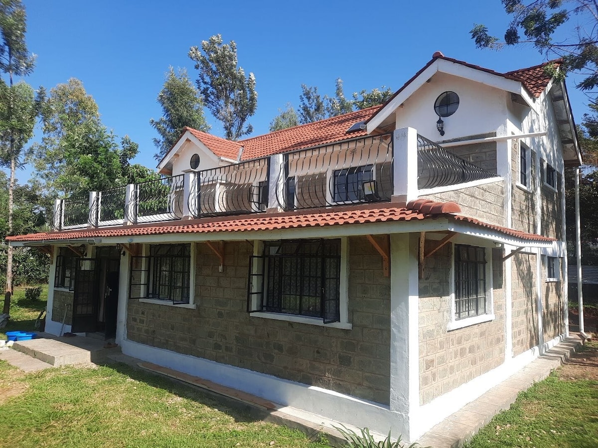 The exterior of a two-story stone house is displayed, featuring a terracotta roof and a spacious terrace with a railing. The architecture incorporates large windows and a welcoming entrance. Lush greenery surrounds the house, highlighting a tranquil atmosphere.