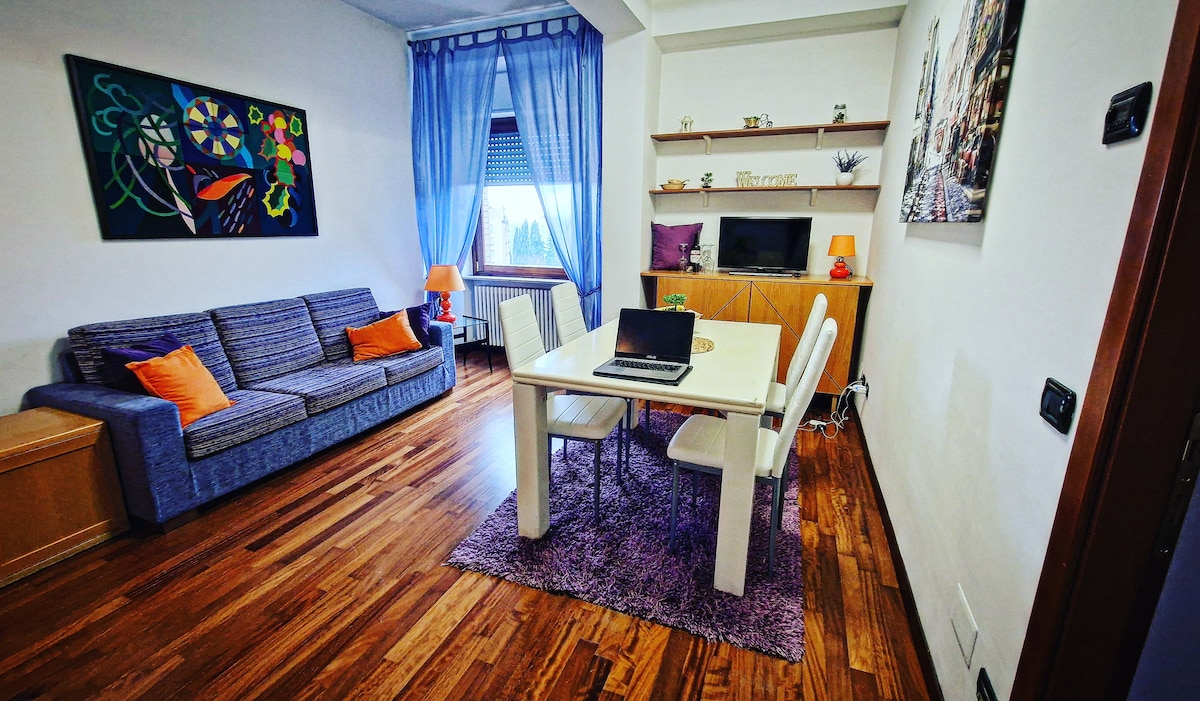A living room is displayed with a blue sofa adorned with orange cushions. A light-colored dining table surrounded by four chairs is positioned on a plush rug. A TV is placed on a wooden cabinet against the wall, complemented by shelves featuring decorative items.