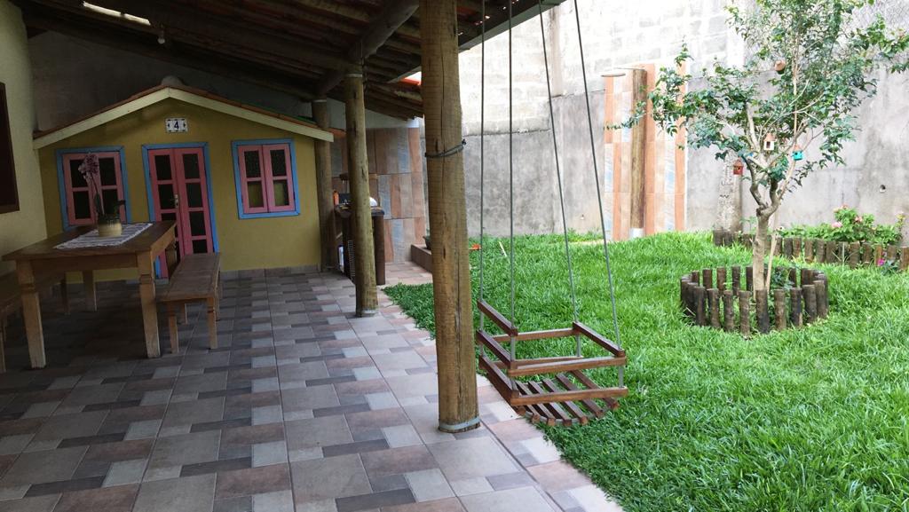 An outdoor area features a grassy lawn surrounded by a wooden fence. A small playhouse with pink windows is visible, next to a wooden picnic table. A swing hangs from a covered patio, adding a playful element to the space.