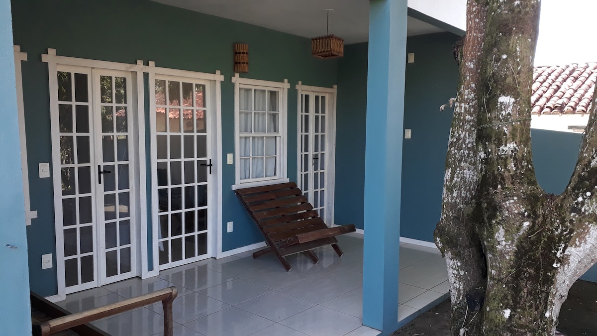 An outdoor seating area features large glass doors leading to an inviting entrance. A wooden lounge chair is positioned near a decorative tree, and the floor is tiled with light-colored tiles. The walls are painted in calming shades of blue and white.