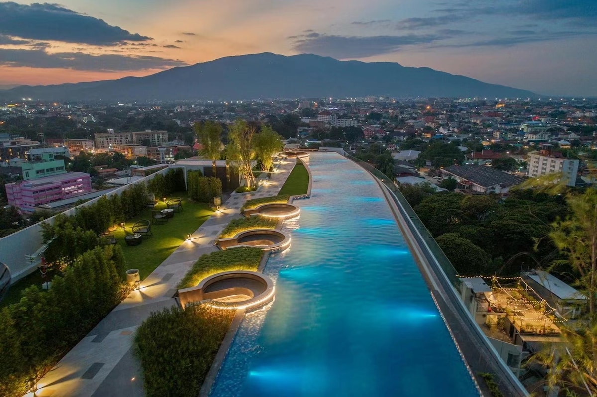 An expansive rooftop infinity pool is visible, surrounded by landscaped greenery. Soft lighting outlines the pool's contours, creating a serene environment. The distant silhouette of mountains contrasts with the cityscape, providing a scenic backdrop at dusk.