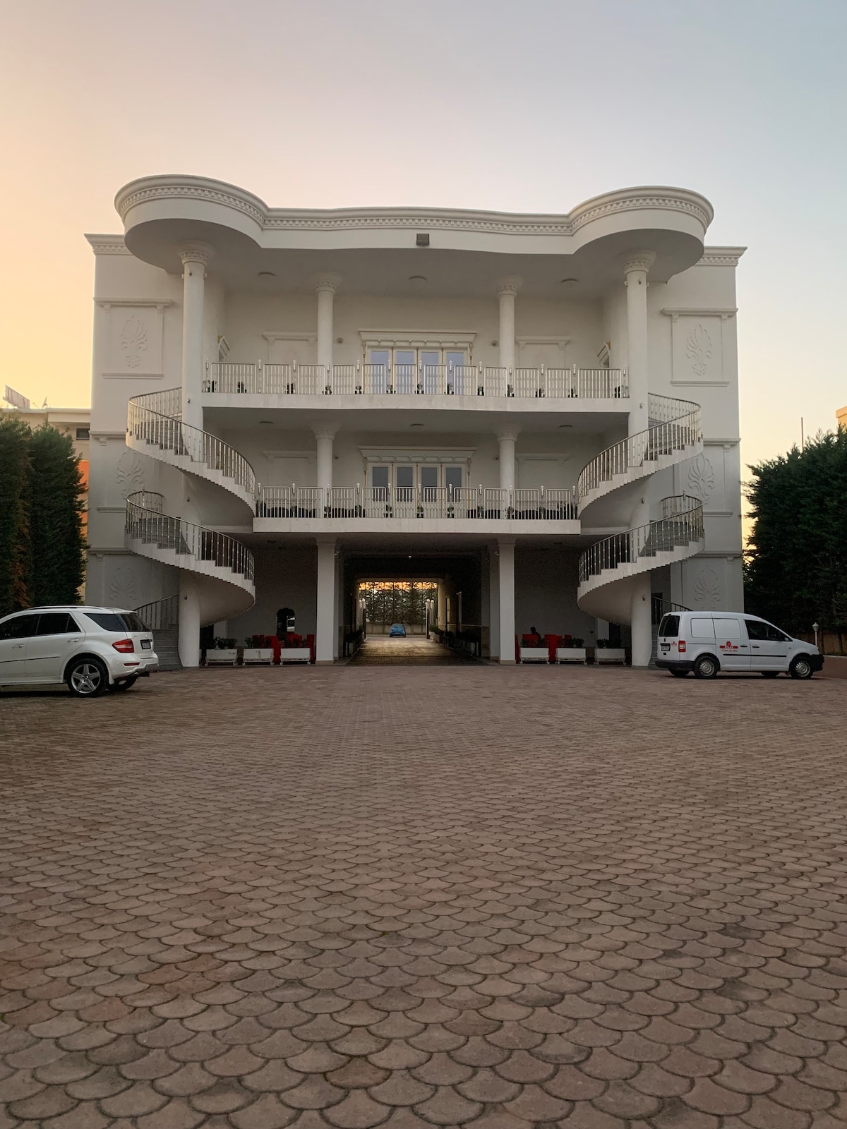 A large, two-story building features elegant architectural details and white columns. A spacious driveway is lined with neatly trimmed shrubs. Two vehicles are parked in front, and a curved staircase leads to the upper level, offering an inviting entrance.