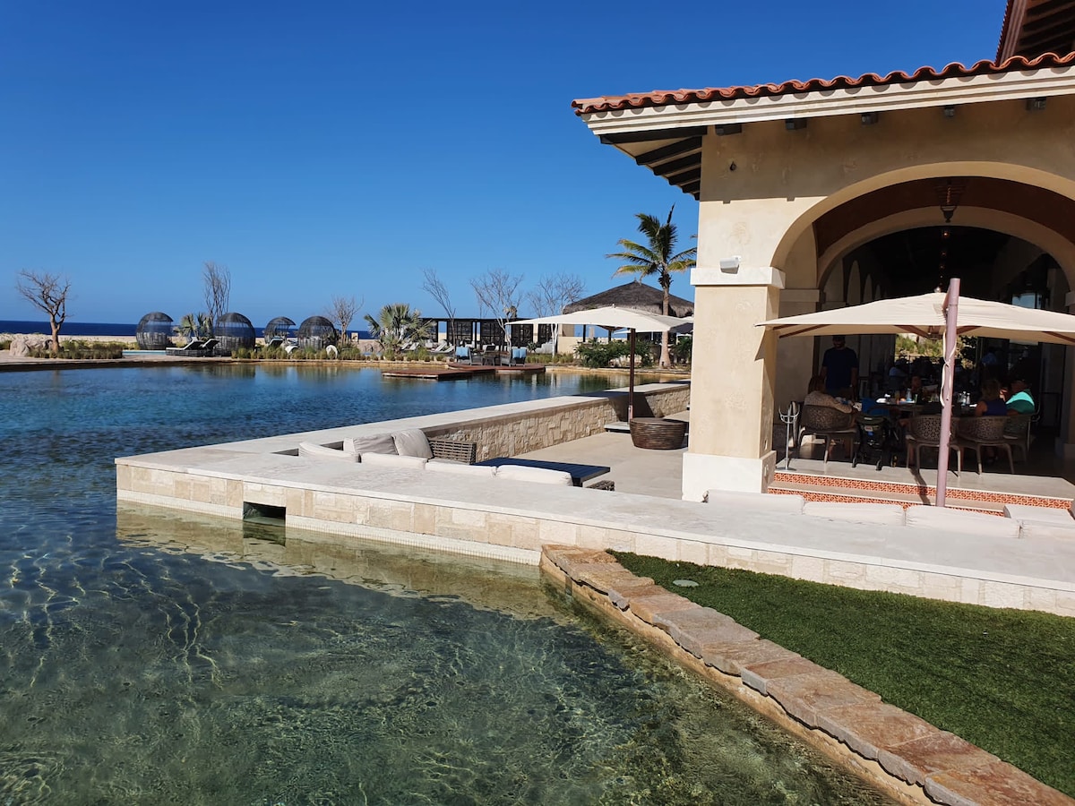 A serene outdoor space features a tranquil lagoon-like area with clear water gently lapping against a stone terrace. Comfortable seating is arranged beneath a large umbrella, set against a backdrop of palm trees and a clear blue sky.