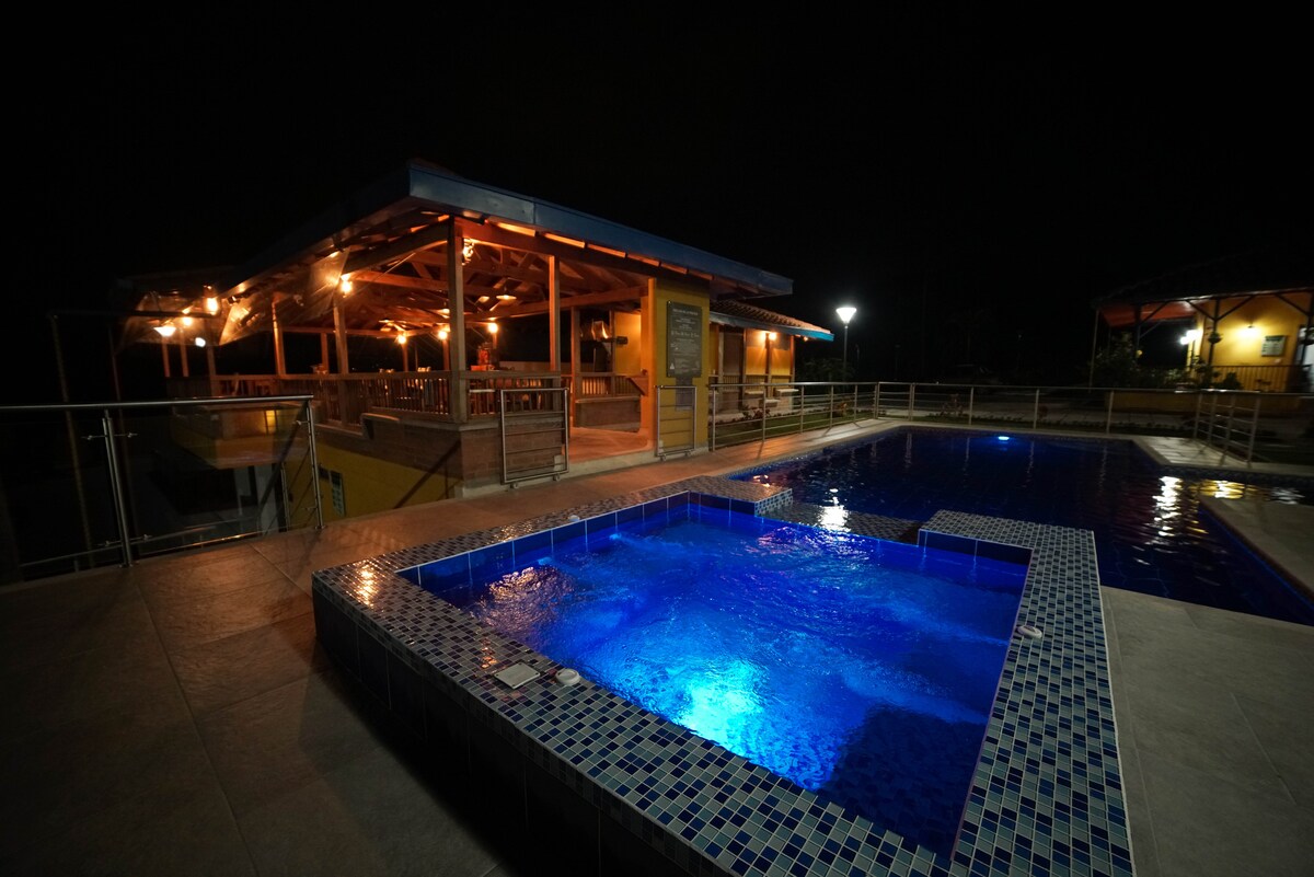 A well-lit outdoor space features a large pool with turquoise lighting, creating a serene ambiance at night. A dining area under a roof is visible, surrounded by wooden railings and additional lighting, while nearby traditional-style accommodations are softly illuminated.