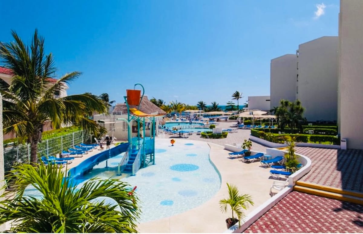 A spacious outdoor area features multiple swimming pools surrounded by loungers and palm trees. A water slide stands prominently in the children's section, while nearby shaded seating areas offer relaxation. Lush greenery lines the pool area, enhancing the inviting environment.