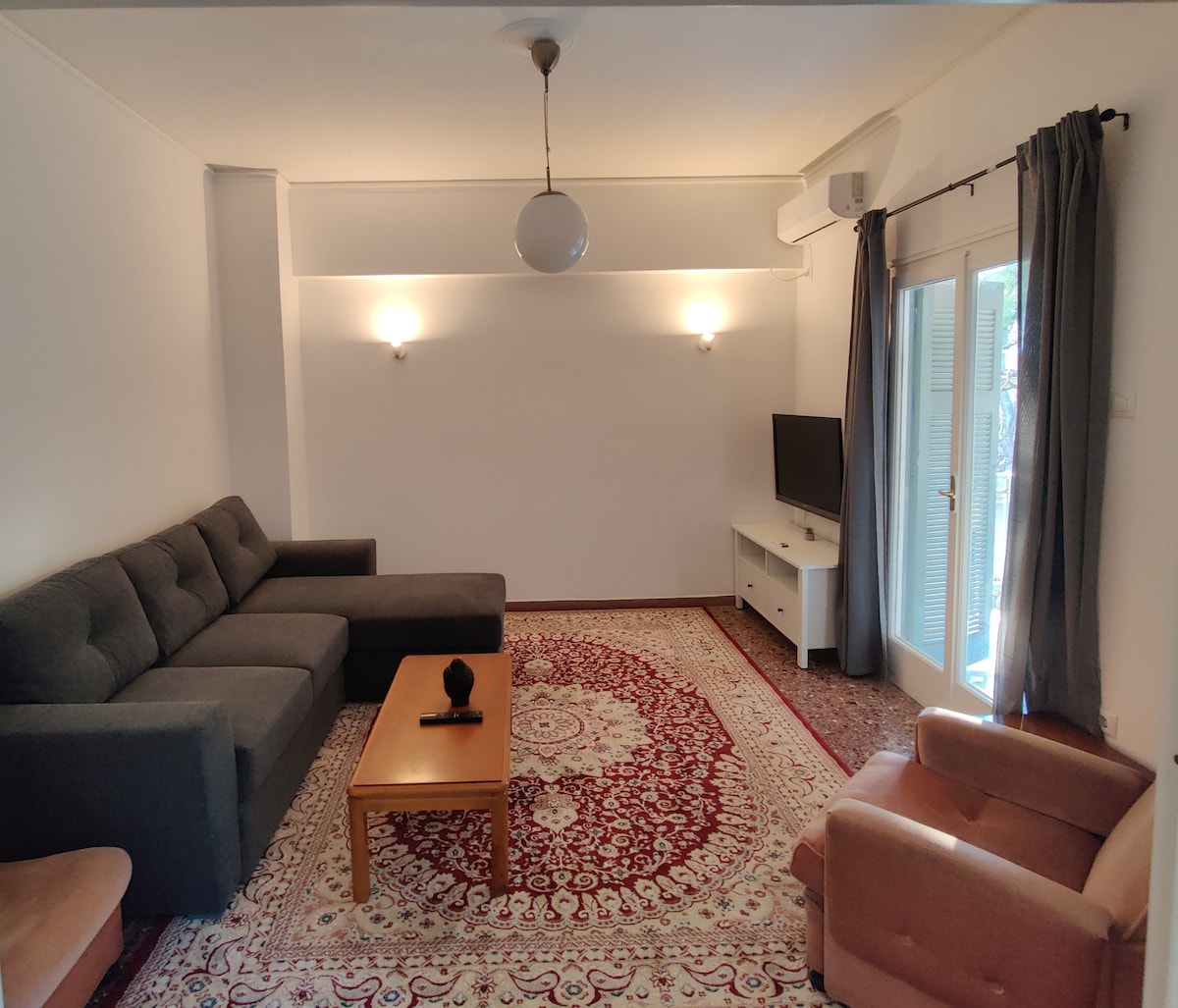 A spacious living area is furnished with a large gray sectional sofa and two armchairs. A coffee table sits in front of the sofa, and a television is mounted on the wall. Natural light enters through a set of double doors leading to a balcony.