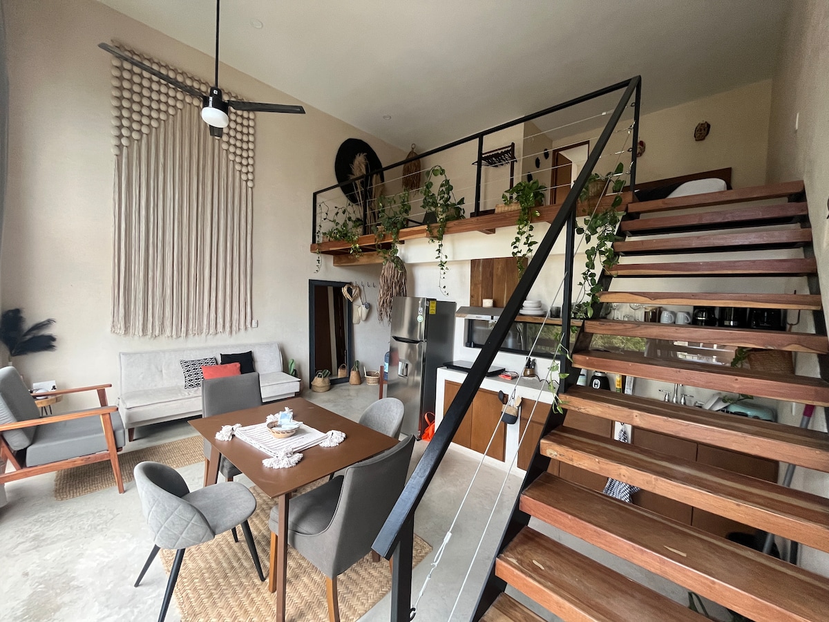 A spacious living area features a dining table set for four and a cozy seating arrangement. The staircase with wooden steps leads to the upper level. Natural light filters through a large window, illuminating the plants adorning the room and adding a touch of greenery.