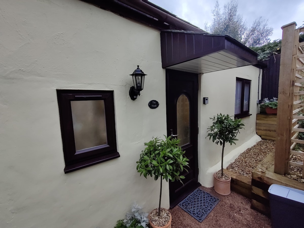 The exterior of the mini-cottage showcases a quaint entrance with a wooden door framed by decorative plants in pots. A vintage-style lantern hangs beside the door, adding a touch of charm. The smooth, light-colored walls and small windows contribute to the inviting look.