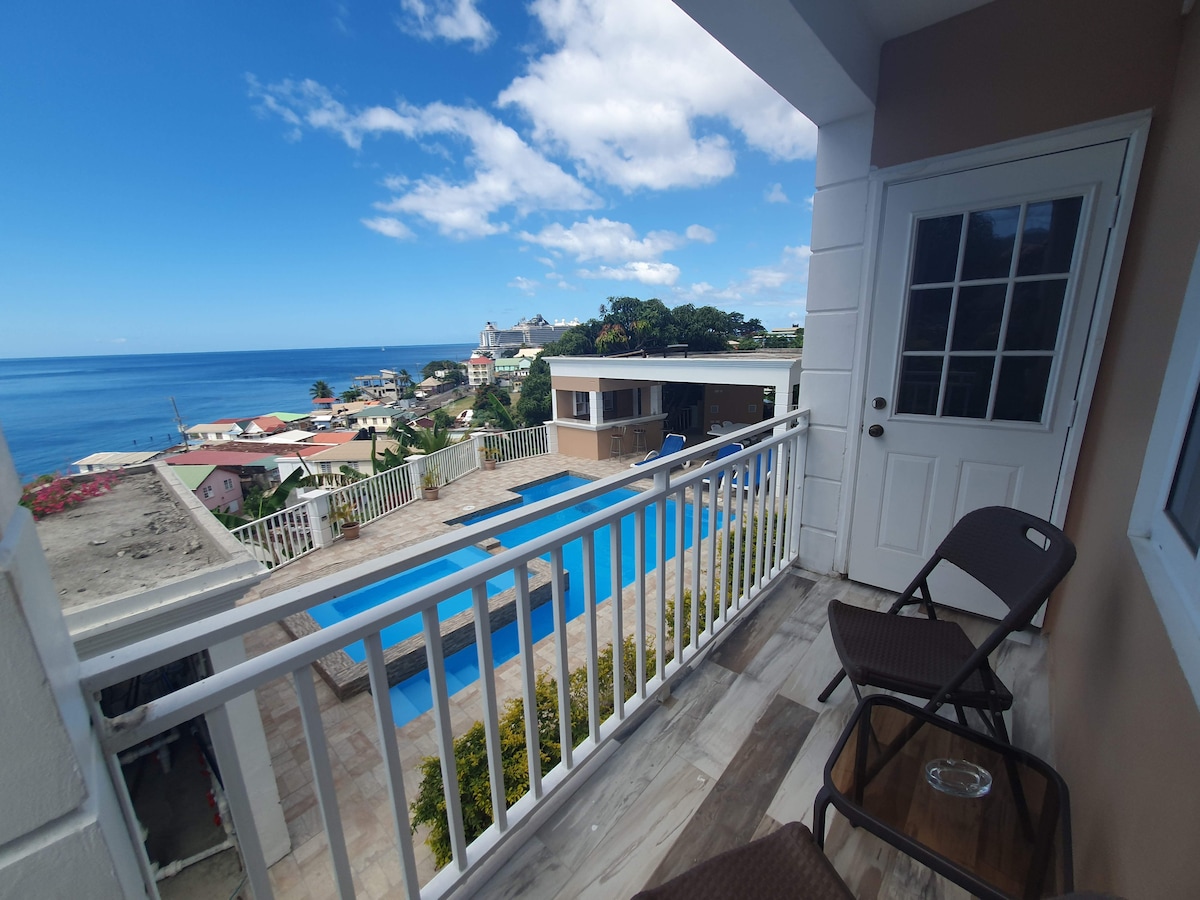 A serene balcony view showcases a spacious pool alongside a vibrant coastal landscape. Two chairs are positioned for relaxation, with the ocean stretching into the horizon. The bright sky is adorned with a few clouds, enhancing the tranquil atmosphere.