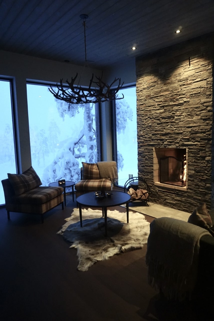 A cozy lounge area features a stone fireplace and two patterned armchairs positioned around a circular coffee table. Large windows provide views of a snowy landscape, and a soft rug lies on the floor. Warm light is emitted from strategically placed ceiling fixtures.