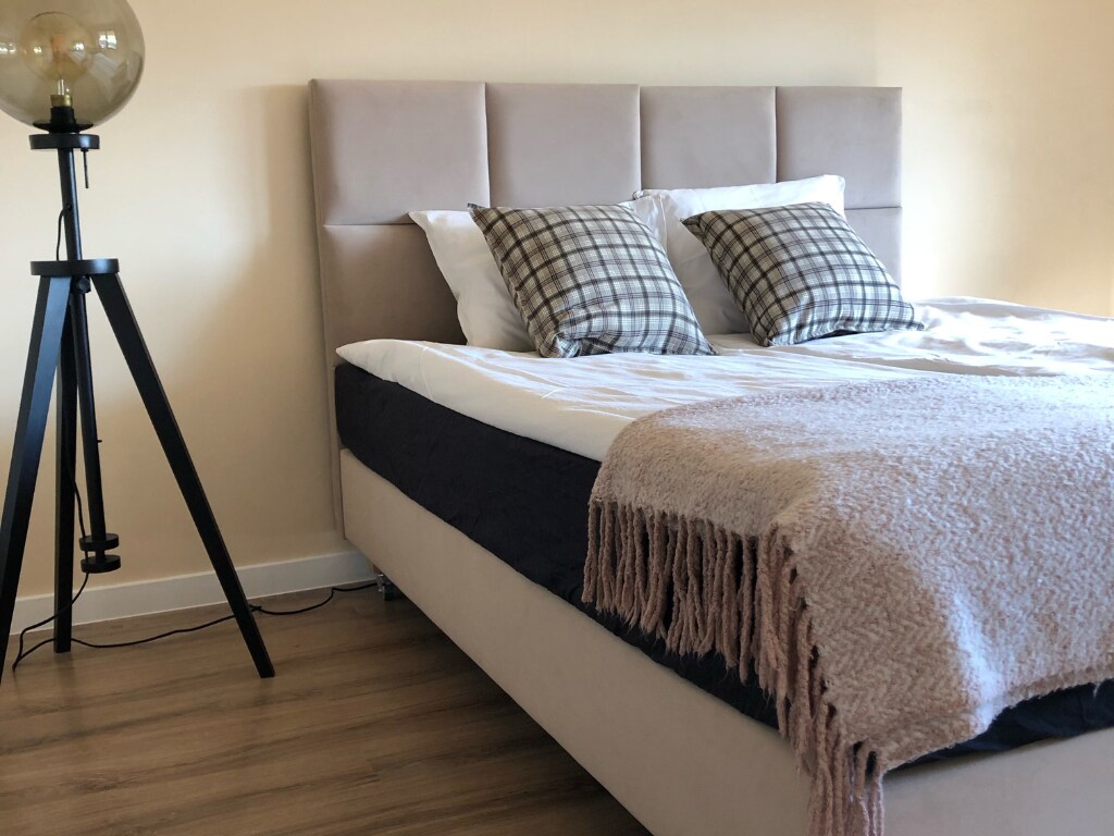 A spacious bed is adorned with decorative pillows and a soft throw blanket. Next to the bed, a modern floor lamp stands on a sleek tripod base. Natural light fills the room, highlighting the warm tones of the walls and wooden flooring.