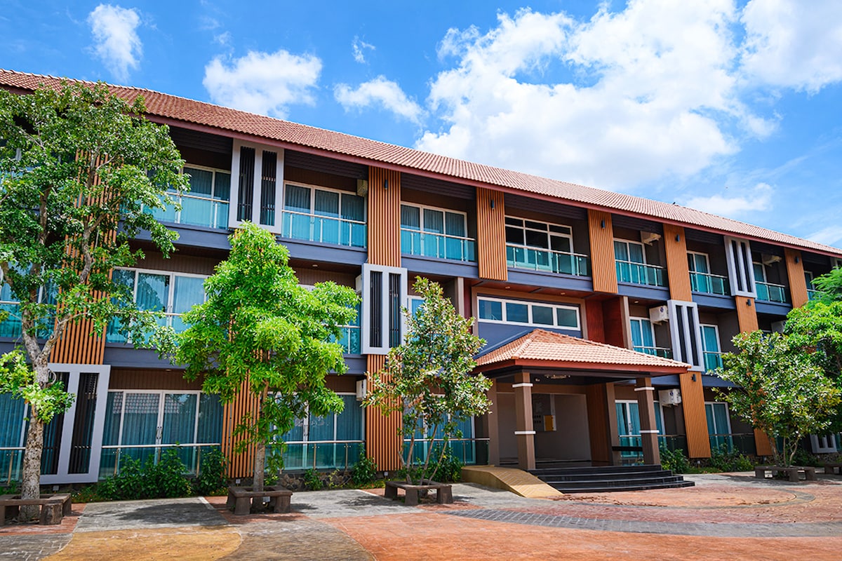 A modern resort building is showcased with a combination of warm wooden and vibrant blue elements. Large glass windows provide ample natural light, while surrounding trees enhance the outdoor environment. A covered entrance leads to the main lobby area, inviting guests to enter.
