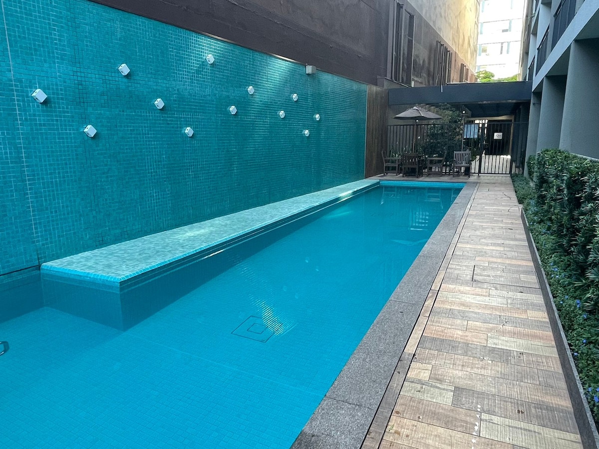 A sleek pool area is surrounded by modern architecture, with water features integrating into the wall. The pool is lined with a textured tiled edge, and a wooden pathway leads to seating areas, providing a calm environment for relaxation.