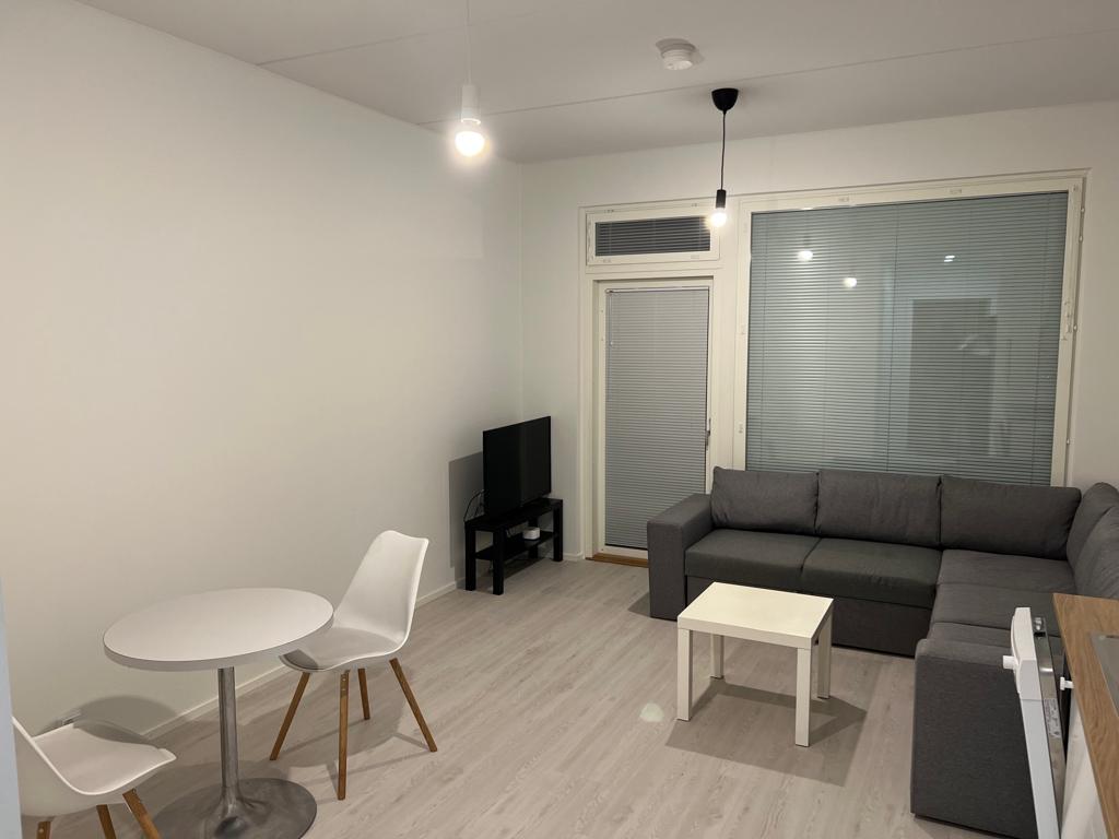 A furnished living space includes a gray sectional sofa and a small, white table surrounded by four chairs. A television is positioned on a stand against the wall. Large windows with blinds allow natural light to enter the room.