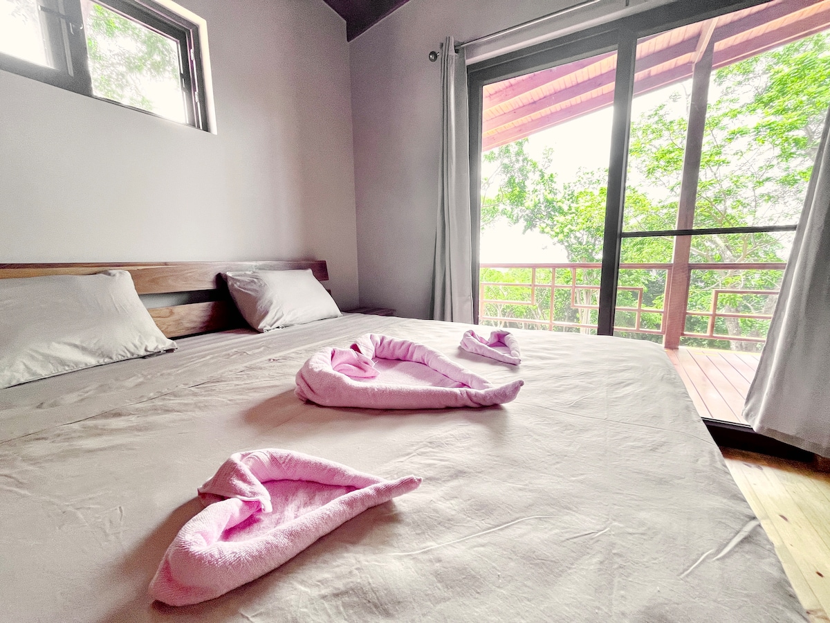 A spacious bed is adorned with neatly arranged pink towels shaped like swans. Large glass doors provide access to a wooden terrace, allowing natural light to fill the room and showcasing views of the surrounding greenery.