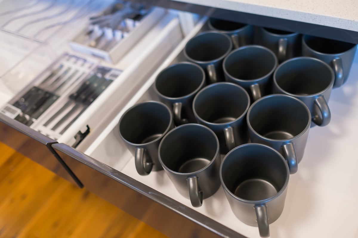 A modern kitchen drawer is opened, displaying twelve black mugs neatly arranged alongside a set of cutlery. The drawer features a clean design, with a contemporary countertop visible above.