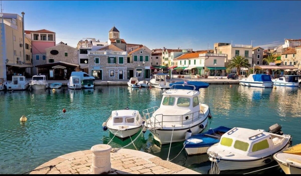 A serene waterfront scene features several boats docked in clear waters, surrounded by charming buildings with varying architectural styles. The harbor is lined with palm trees, and small fishing vessels are visible alongside larger boats, reflecting a tranquil atmosphere.