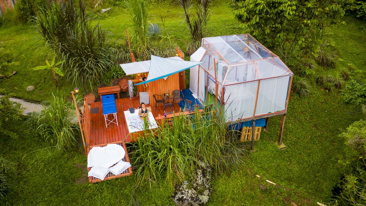 A cozy glamping structure is nestled in greenery, featuring a wooden deck with seating areas. A transparent enclosure provides a bright space, complemented by a shaded area with colorful textiles. Lush grass and plants surround the setup, enhancing the connection to nature.
