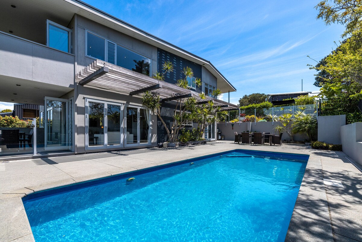 A modern home is showcased with a private swimming pool surrounded by a spacious patio. The backyard features greenery and outdoor dining spaces, with large glass doors providing a view into the interior. The home's clean lines and elegant design enhance the outdoor area.
