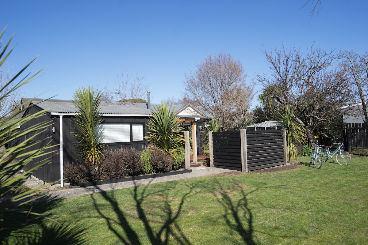 A single-story dark wooden building is set within a landscaped yard, featuring low-maintenance plants and trees. A gravel path leads to the entrance, while two bicycles are parked nearby, suggesting leisurely exploration of the surrounding area.
