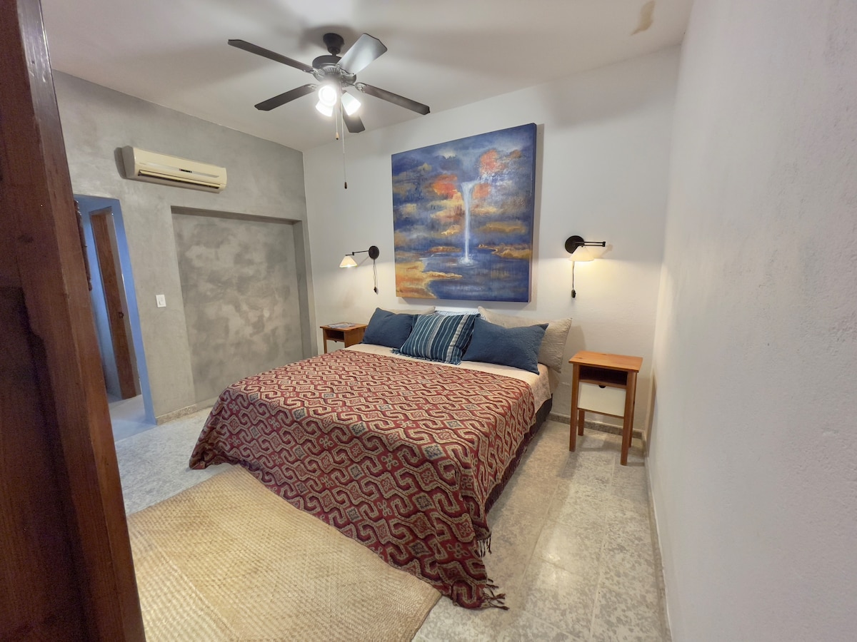 A spacious bedroom features a king size bed with a patterned coverlet and blue accent pillows. Soft ambient lighting is provided by wall sconces beside the bed. A ceiling fan hangs above, and a large piece of wall art adds color to the neutral-toned room.