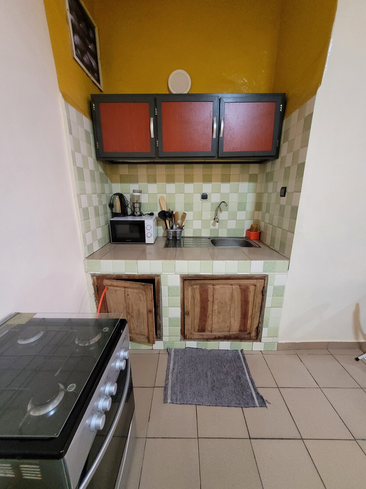 A compact kitchen area features wooden cabinets with a contrasting green and white tiled backsplash. A sink and countertop are situated below the cabinets, alongside a microwave and cooking utensils. A gas stove is positioned nearby, adding functionality to the space.