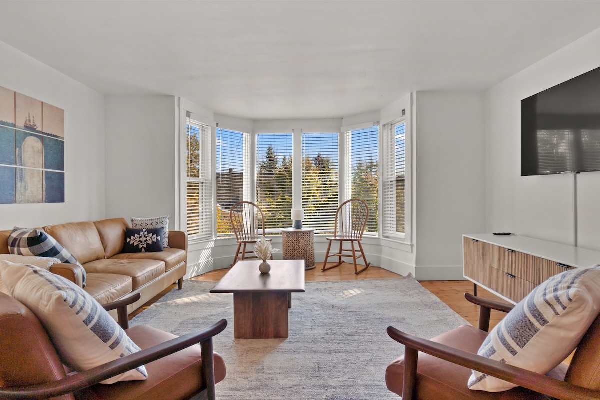 A sunlit living area features large bay windows with blinds, providing a view of surrounding trees. A leather sofa and two wooden chairs are arranged around a central coffee table on a soft area rug, complemented by decorative pillows.