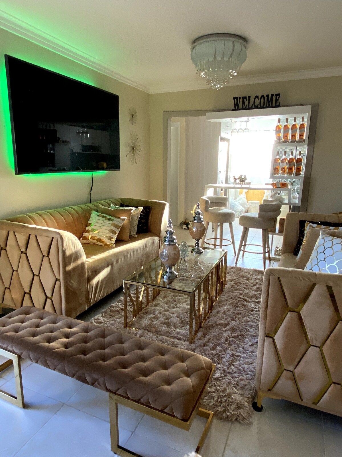 A welcoming living area features plush, gold-accented sofas arranged around a glass coffee table, adorned with decorative vases. A bar area is visible in the background, with shelves displaying a variety of bottles. Soft lighting casts a gentle glow throughout the space.