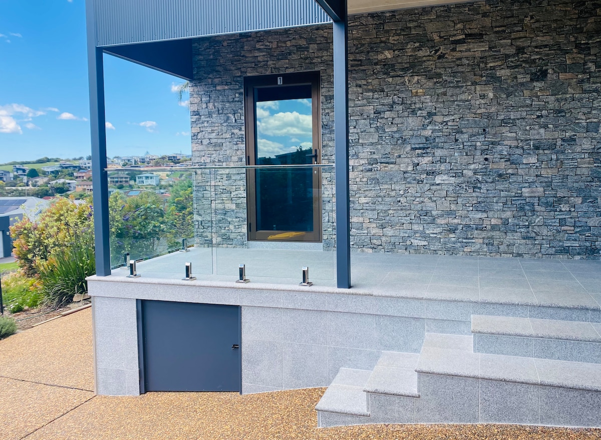 An outdoor space features a large patio area with stone walls and a modern glass railing. Steps lead down to a lower level, with a small door integrated into the design. Lush greenery and a view of the surrounding landscape are visible in the background.