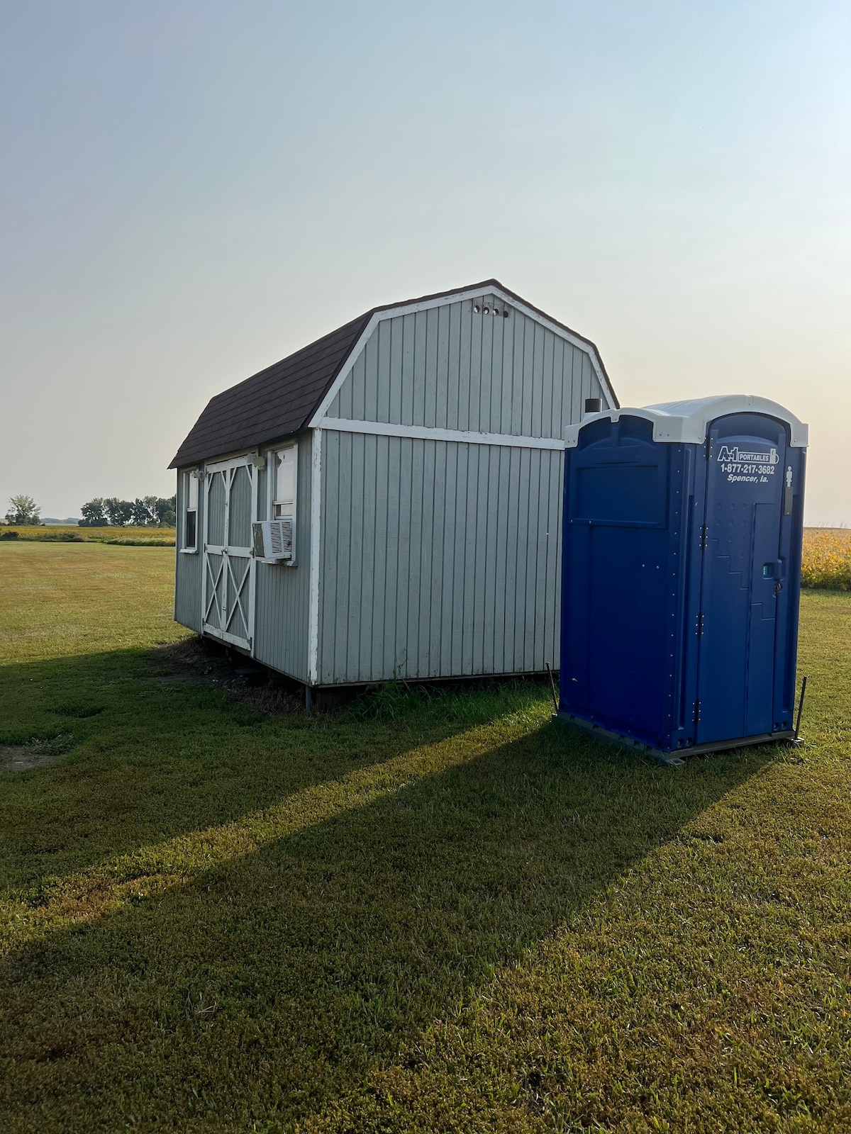 A small cabin with a gabled roof is positioned in an open grassy area, accompanied by a blue portable toilet nearby. An air conditioning unit is visible on the cabin's side, with ample space surrounding the structure for outdoor activities.