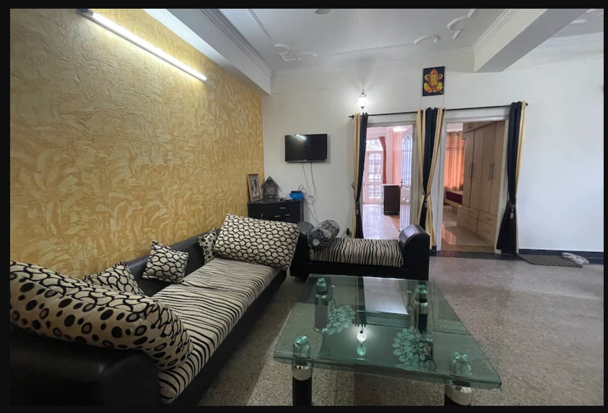 A stylish living area features a modern black sofa adorned with patterned cushions. A glass coffee table sits in the center, reflecting light. A wall-mounted television is visible, while soft lighting illuminates the space, creating an inviting atmosphere that connects to the adjoining rooms.