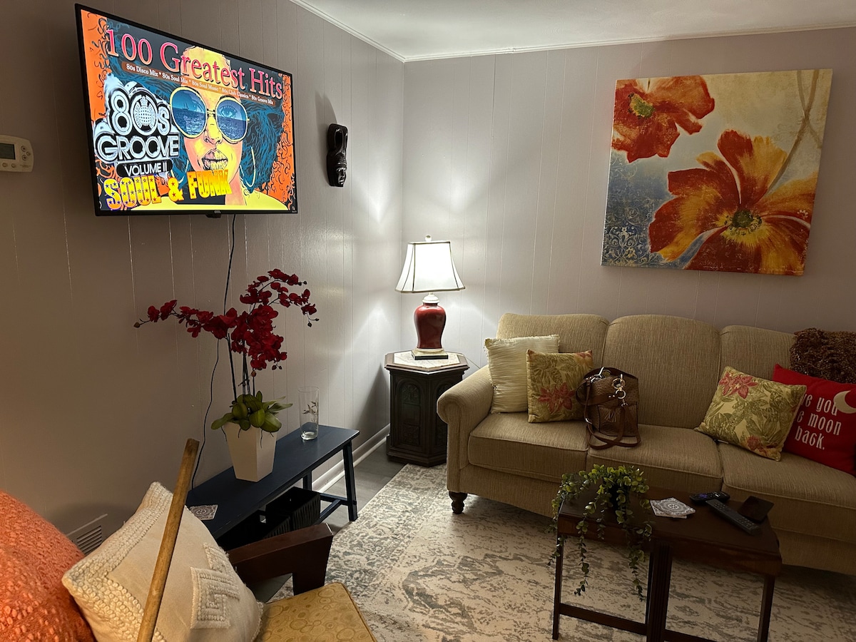 A cozy living area features a light-colored sofa and decorative cushions. A flat-screen TV displays music content, while a lamp provides soft lighting. An ornamental plant sits on the coffee table, complemented by a rug that adds texture to the space.