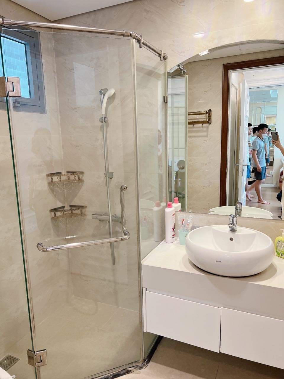 A modern bathroom is depicted, featuring a glass-enclosed shower with a showerhead mounted on the wall. A round sink is positioned on a sleek countertop, accompanied by neatly arranged toiletries. A large mirror reflects the space, enhancing the sense of openness.