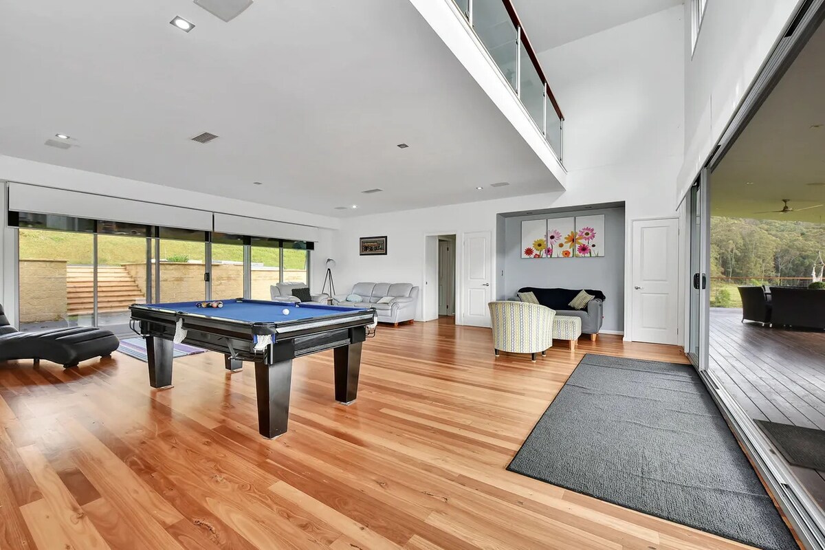 A spacious games room features a billiard table, smooth wooden flooring, and large glass doors that offer views of the outdoors. Relaxation areas are present, with comfortable seating arranged for conversation. Natural light fills the room, enhancing the open and airy ambiance.