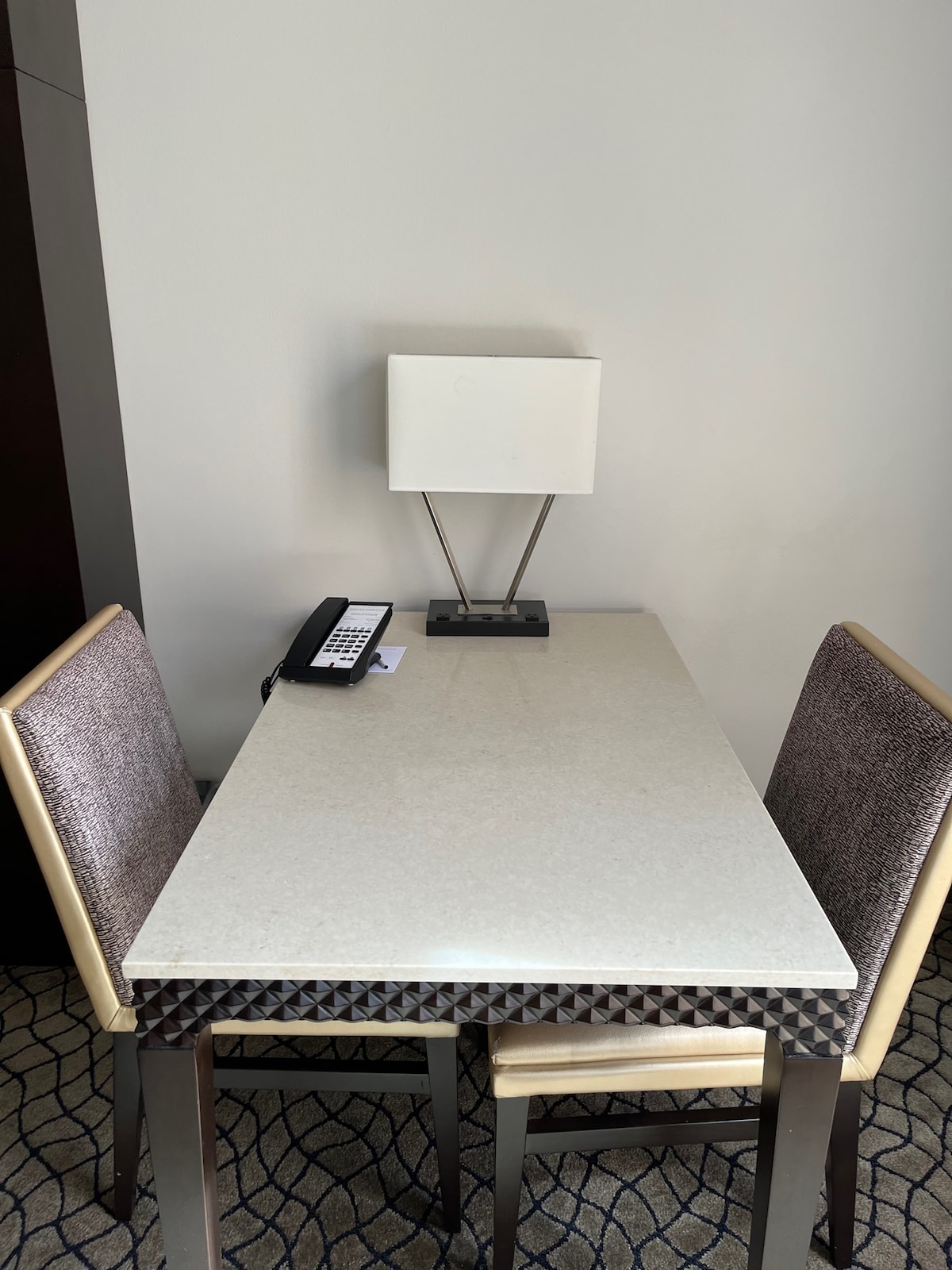 A simple table is paired with two chairs, positioned against a wall. A modern lamp stands on the table, providing light. A telephone is placed nearby, adding practicality to the space.
